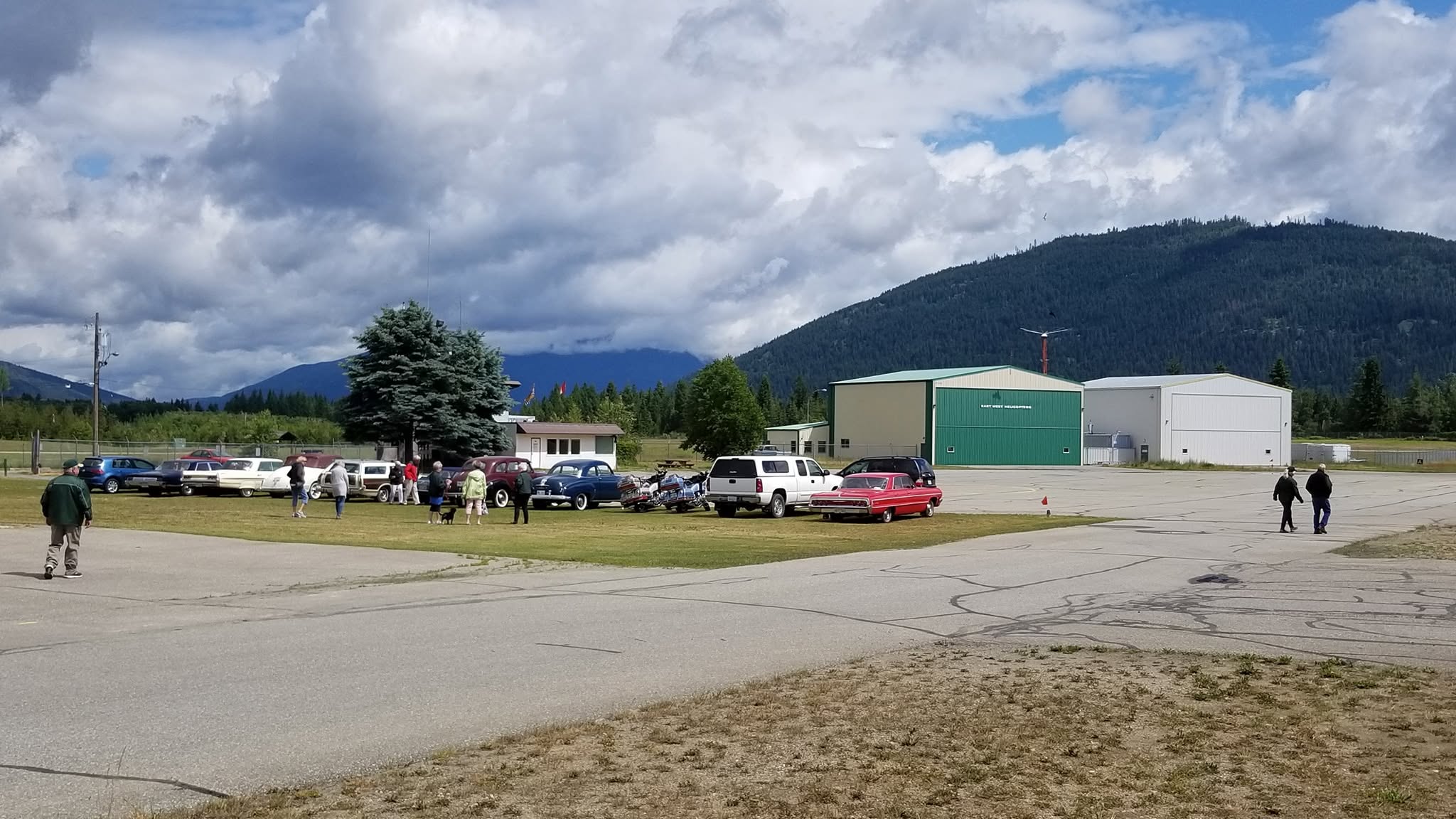 Classic cars and visitors on the grass beside the hangars at the Salmon Arm airport, with forested mountains behind