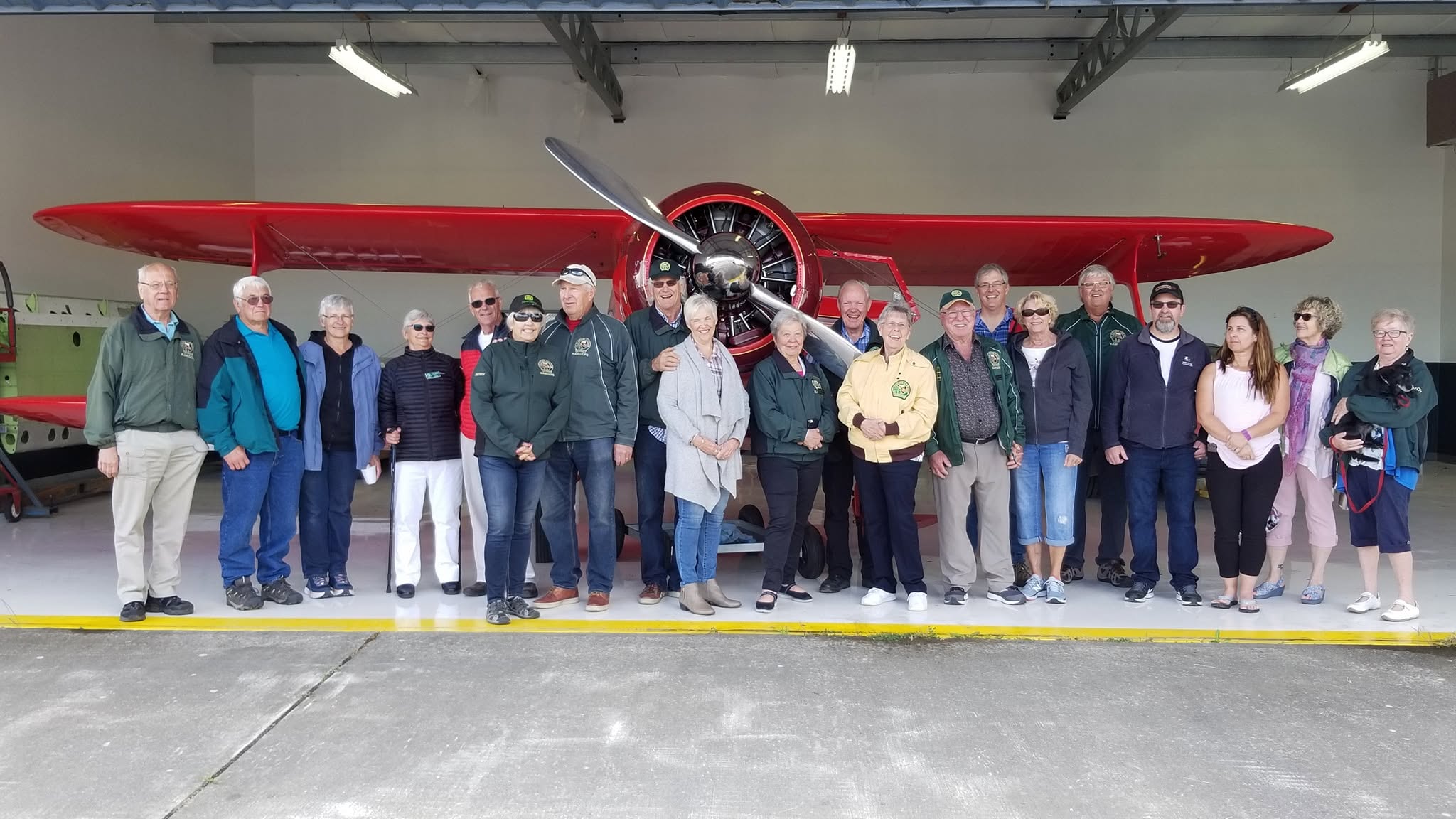 Club members, many in green club jackets, standing in a row in front of a red radial-engine biplane in a hangar